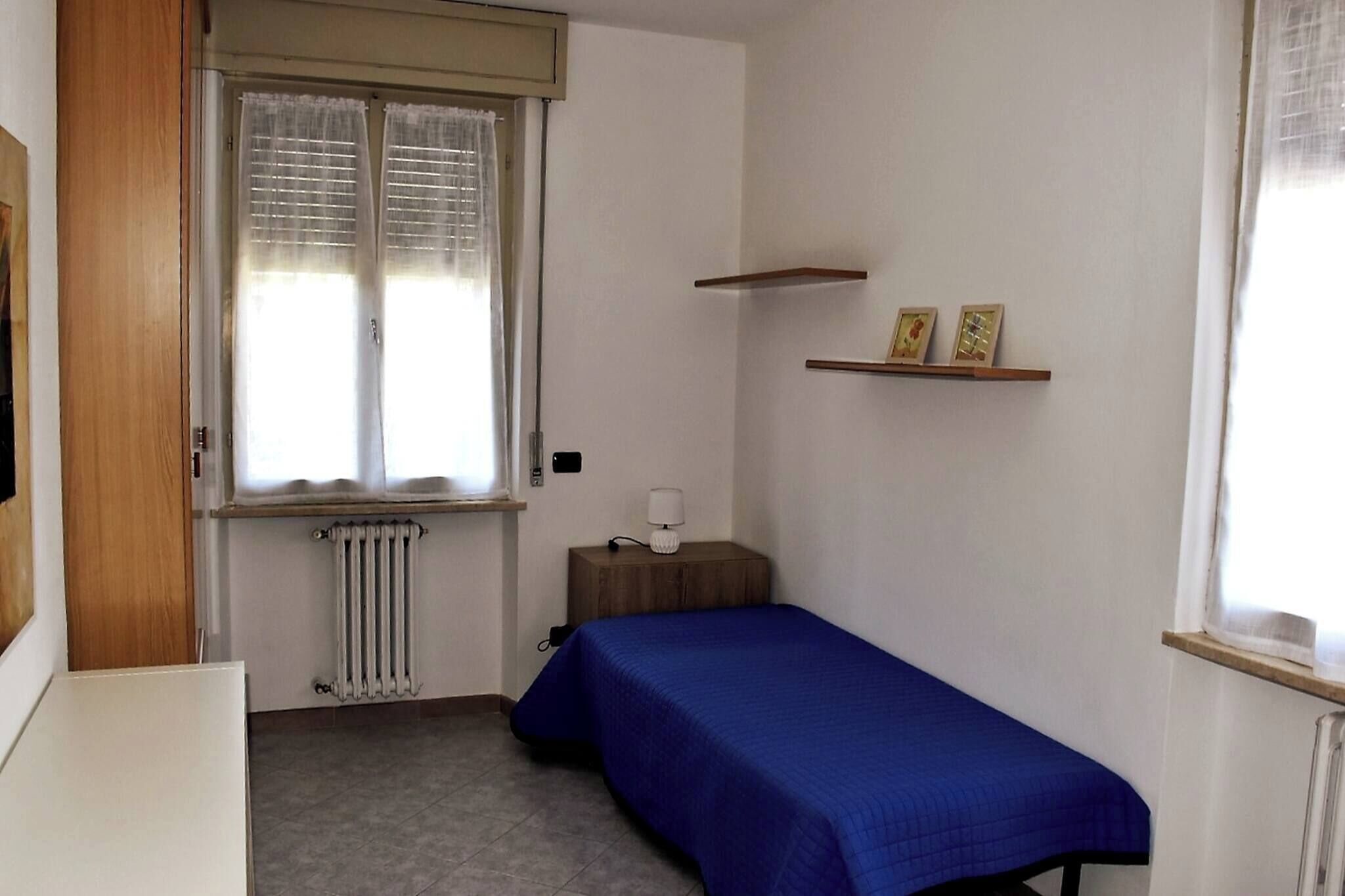 Una habitación pequeña y ordenada con una cama individual, un radiador, una ventana con persianas, una mesita de noche con lámpara y dos marcos de fotos en un estante.