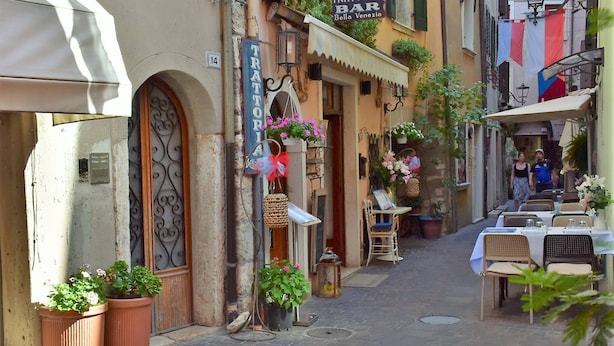 Eine enge Straße mit Außenbestuhlung, einer Bar namens "Trattoria Bella Venezia" und Blumentöpfen.