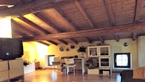 Een ruime woonkamer met een houten plafond, een televisie, een eethoek met een tafel en stoelen, en een keuken met kastjes en een raam.