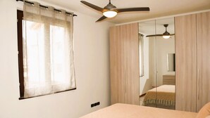 Un dormitori amb llit, ventilador de sostre, finestra amb cortines lleugeres i un mirall que reflecteix l'habitació.