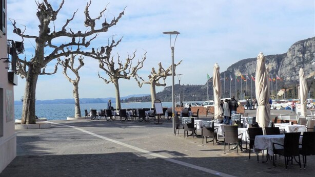An outdoor café with tables and chairs, a view of the sea, and bare trees. - Catullo House in Garda (Garda)