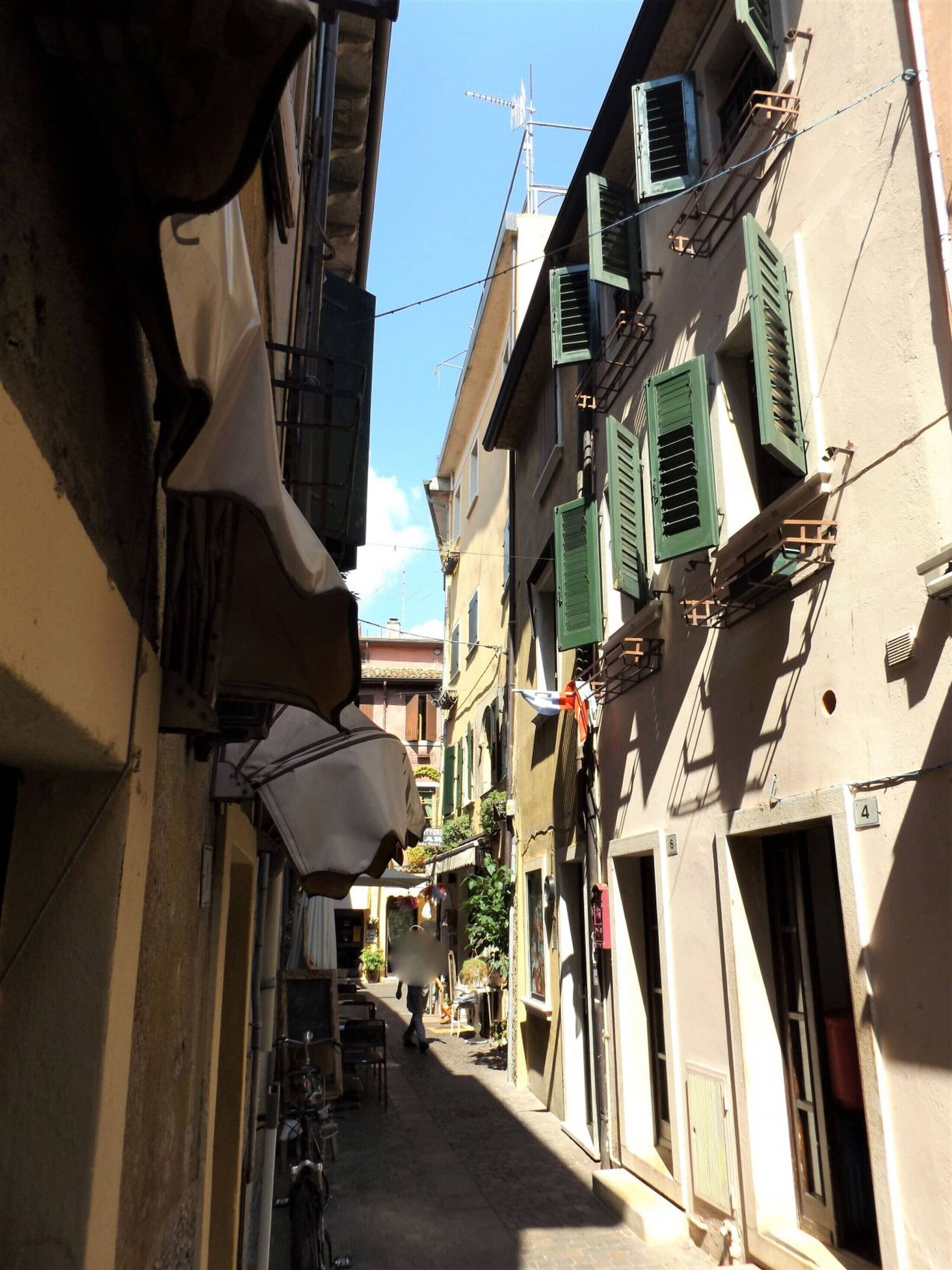 A narrow street with buildings on either side, featuring balconies with green shutters and a clear blue sky above.