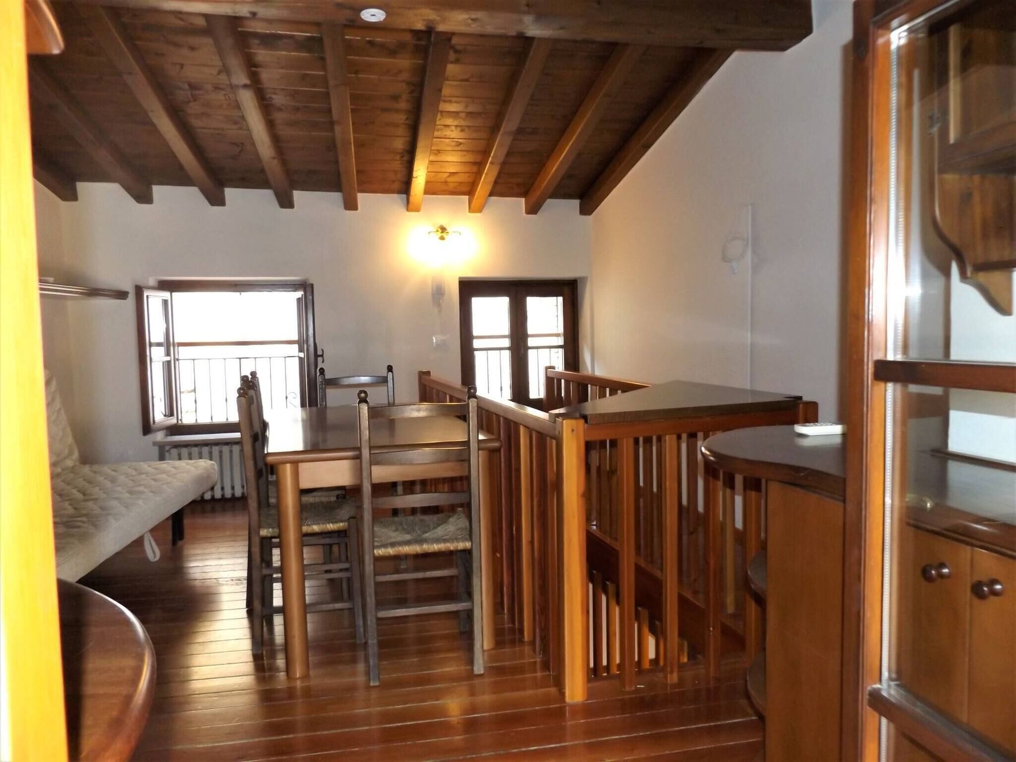 A compact room with a wooden ceiling, a bed, a table, and chairs. There are windows providing natural light.