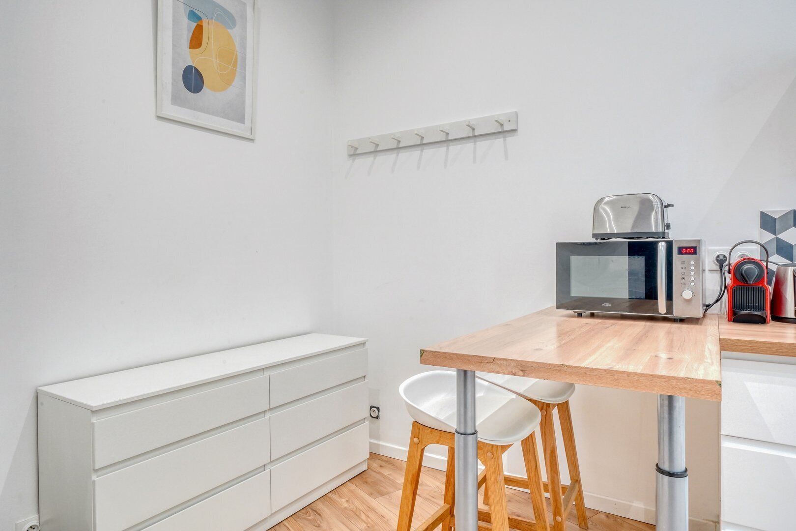 A compact kitchen with a microwave, toaster, and kettle. There is a white cabinet with drawers and a wooden stool.