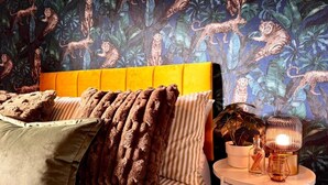 Een kamer met een gestreepte bank, een geel hoofdeinde en behangpapier met luipaardenprint. Er staat een klein rond tafeltje met een lamp en een potplant.