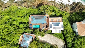 Vista aérea de un complejo turístico con piscina, un edificio independiente con techo solar y exuberante vegetación alrededor de la propiedad.