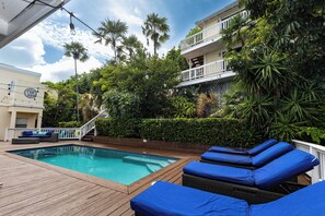 A swimming pool with lounge chairs and a wooden deck, surrounded by lush greenery and a multi-story building with balconies. - The Breeze Villa, newest ocean front boutique oasis (St Thomas)