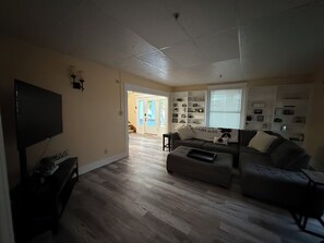 A living room with a flat-screen TV, a sofa, and a coffee table. There is a bookshelf with various items and a door leading to another room.