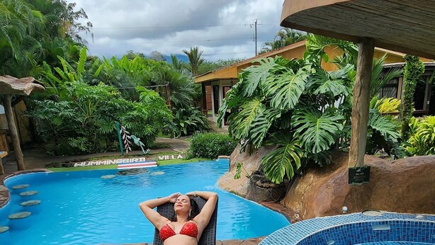 A woman relaxing on a lounge chair by a pool with a tropical garden backdrop. - Hummingbird Villa (La Fortuna)