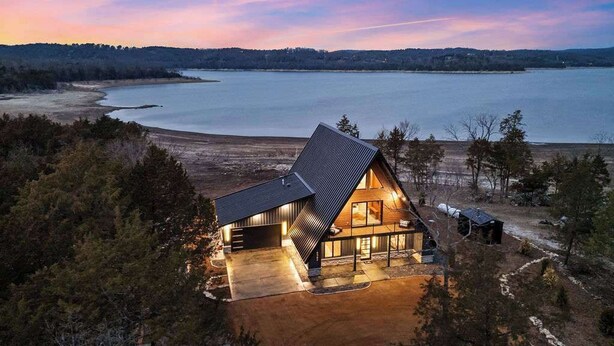 A modern house with a sloped roof, large windows, and a well-lit interior, surrounded by a forest and a lake. - AFrame at Beaver Lake |Hot Tub. Epic Patio & Views (Rogers)
