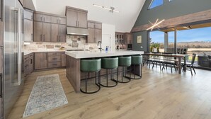 A modern kitchen with a large island, bar stools, and a dining area with a view. - AFrame at Beaver Lake |Hot Tub. Epic Patio & Views (Rogers)