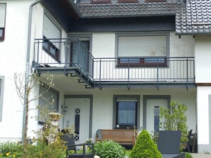 A two-story residential building with a balcony, a small garden, and a wooden bench. - Ferienwohnung in der Vulkaneifel (Winkel)