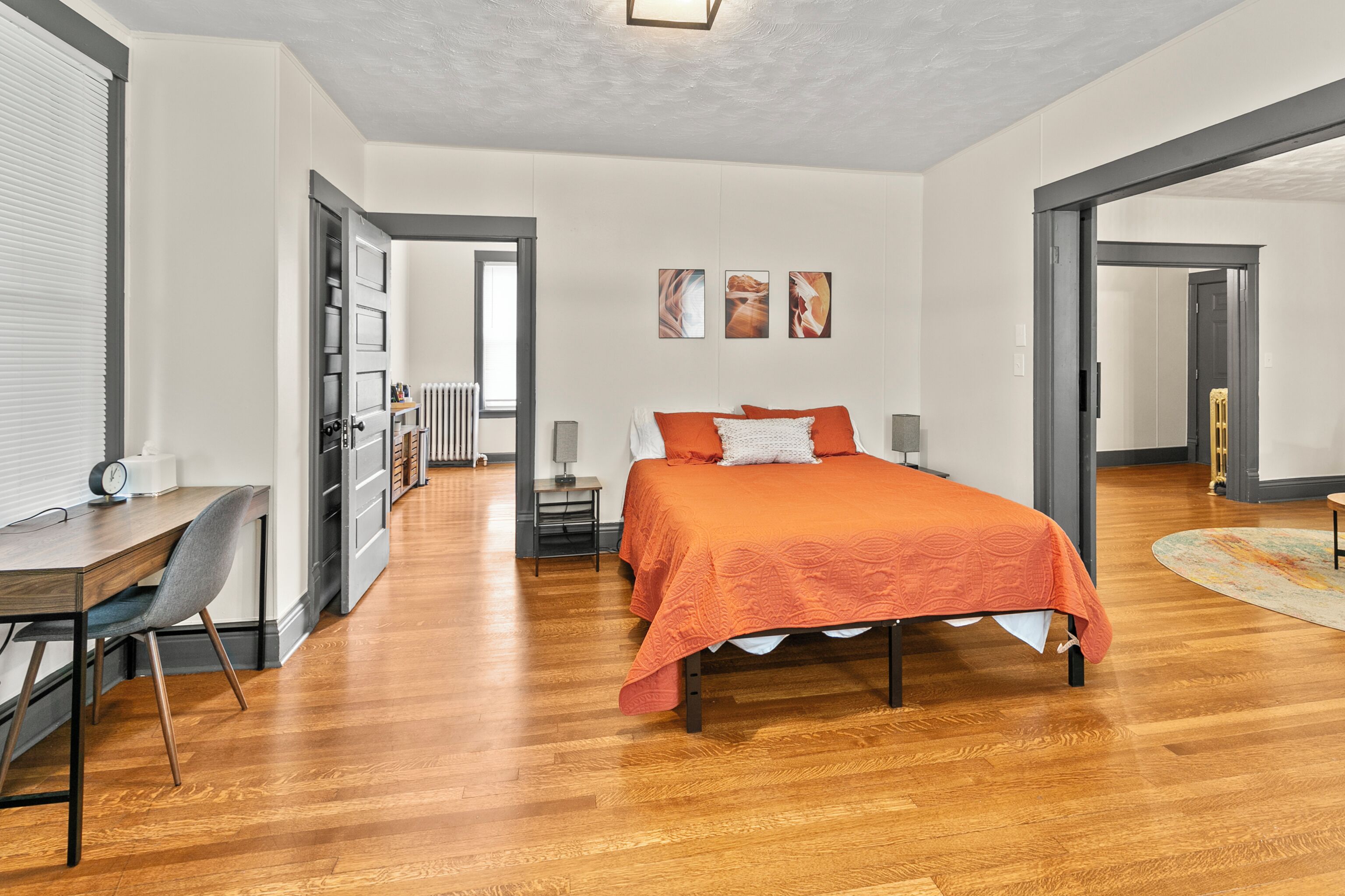 Un dormitorio con piso de madera, una cama con cubrecama naranja, un escritorio con silla y un radiador.