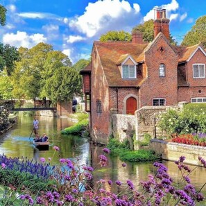 Une maison traditionnelle en briques, avec un canal devant, entourée d’une végétation luxuriante et de fleurs.