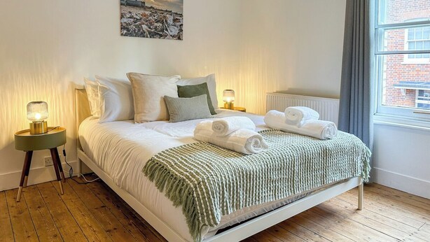 A neatly made bed with white linens and a green throw, two bedside lamps, a painting on the wall, and a window with a view of a brick building. - New | Cosy & Stylish City Centre Home | Sleeps 6 (Canterbury)