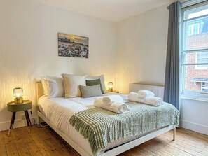 A neatly made bed with white linens and a green throw, two bedside lamps, a painting on the wall, and a window with a view of a brick building.