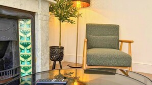 A room with a fireplace, a green armchair, a glass coffee table, a lamp, and a remote control. - New | Cosy & Stylish City Centre Home | Sleeps 6 (Canterbury)