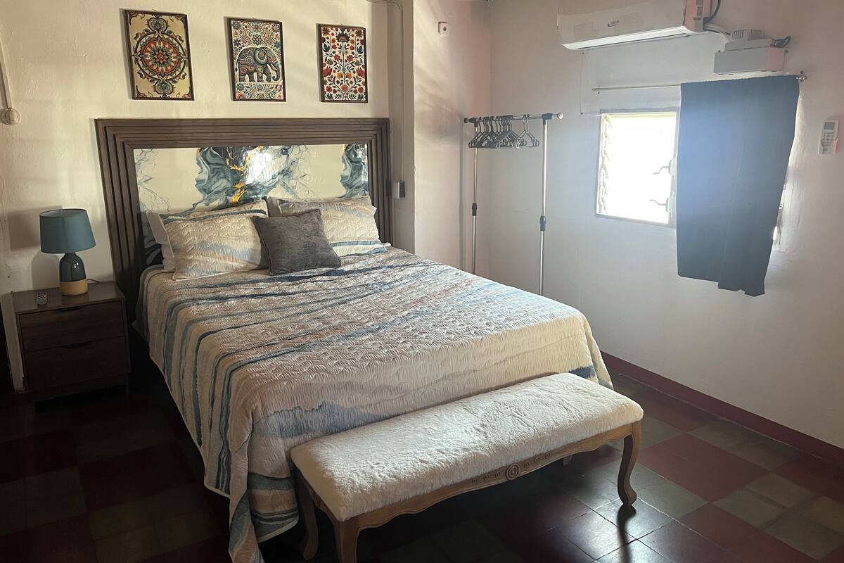 A neatly arranged bedroom with a bed, bedside table, and a bench. There are decorative wall hangings above the bed and a window with a curtain.