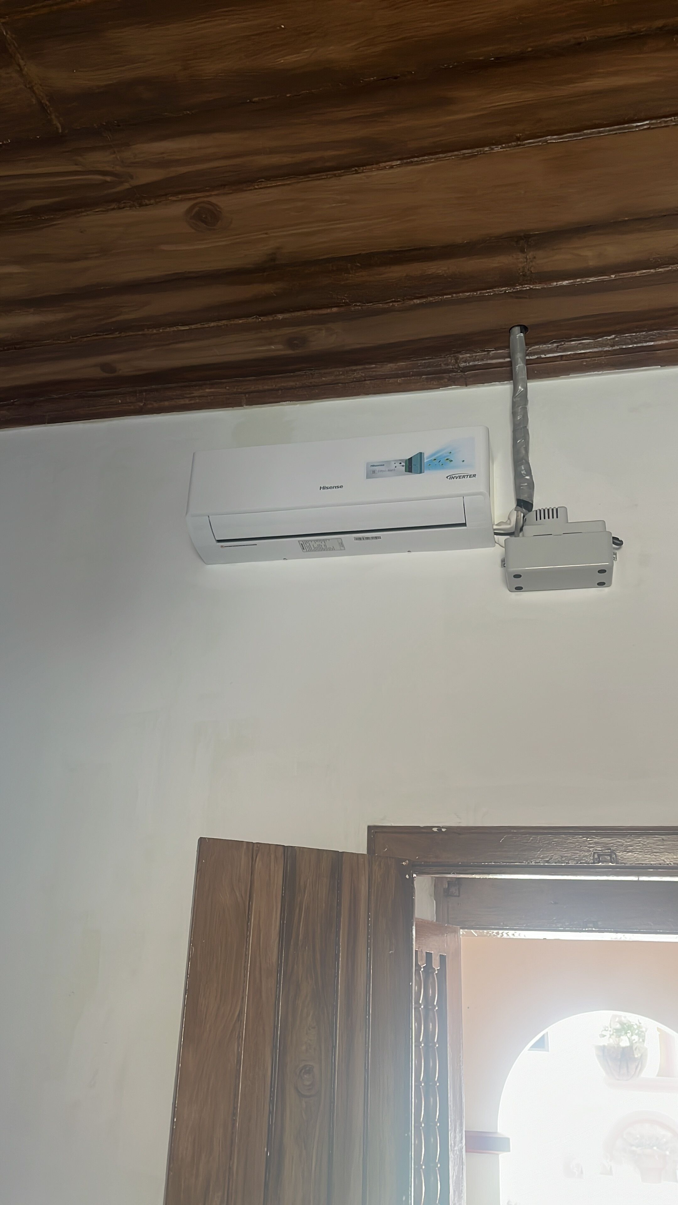 Unitat de aire condicionat penjada al sostre, amb el logo de Hisense, un marc de porta de fusta i un sostre de fusta.