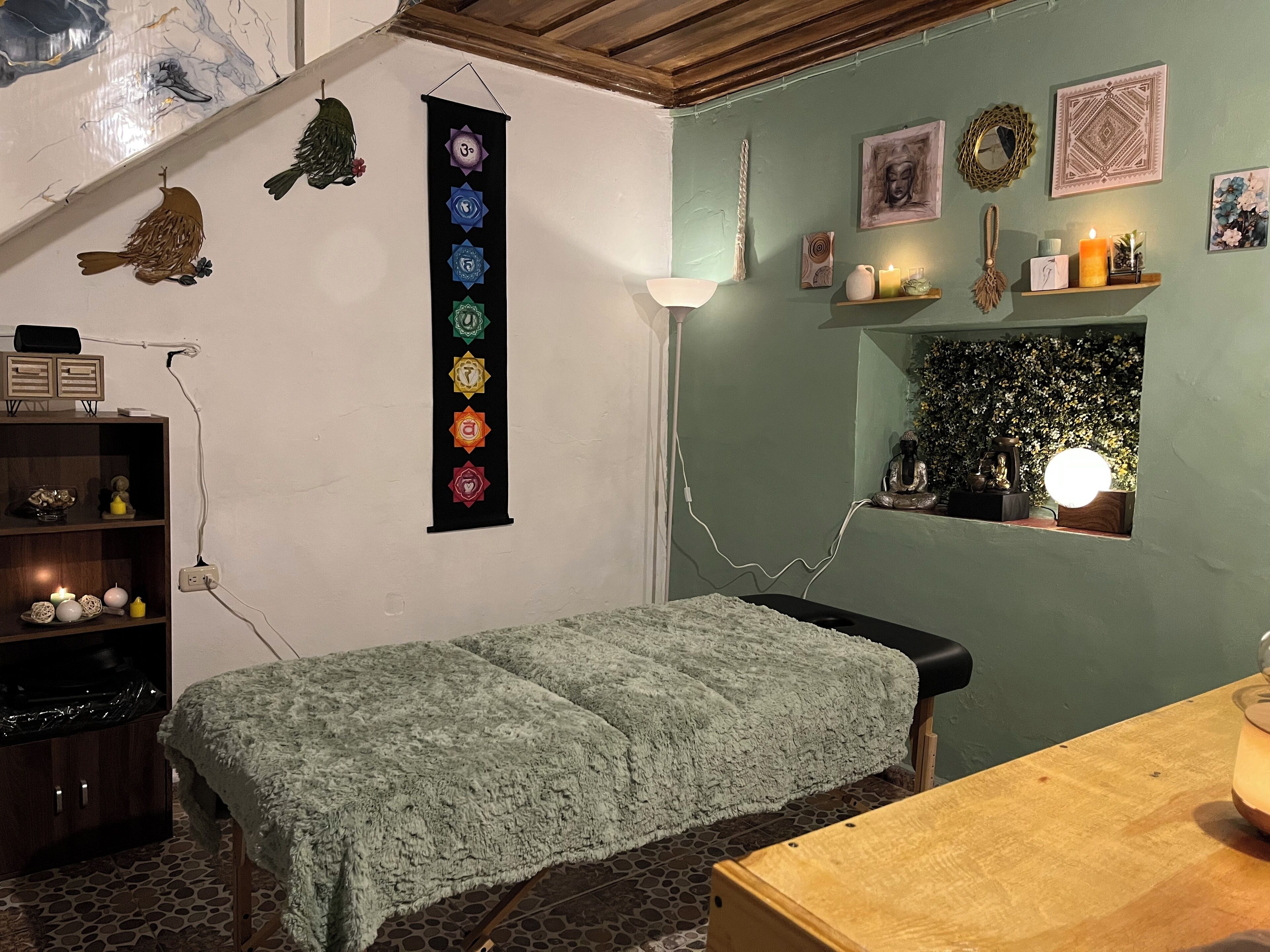 A room with a bed, a small table, a wall-mounted shelf with decorative items, a wall with a vertical emblem, and a window with a Buddha statue.
