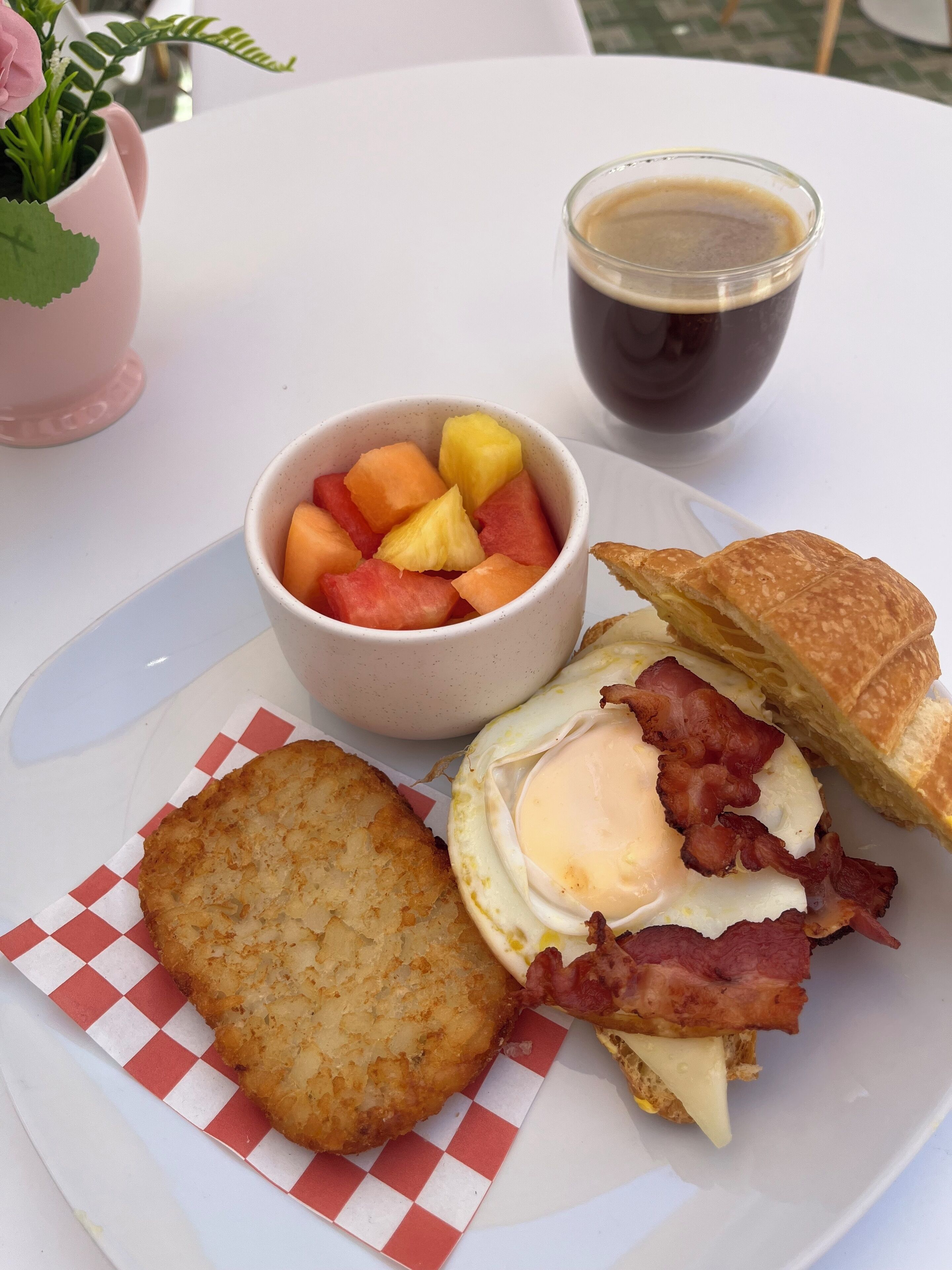 A breakfast plate with hash browns, a sandwich with bacon and egg, a cup of coffee, and a bowl of fruit.