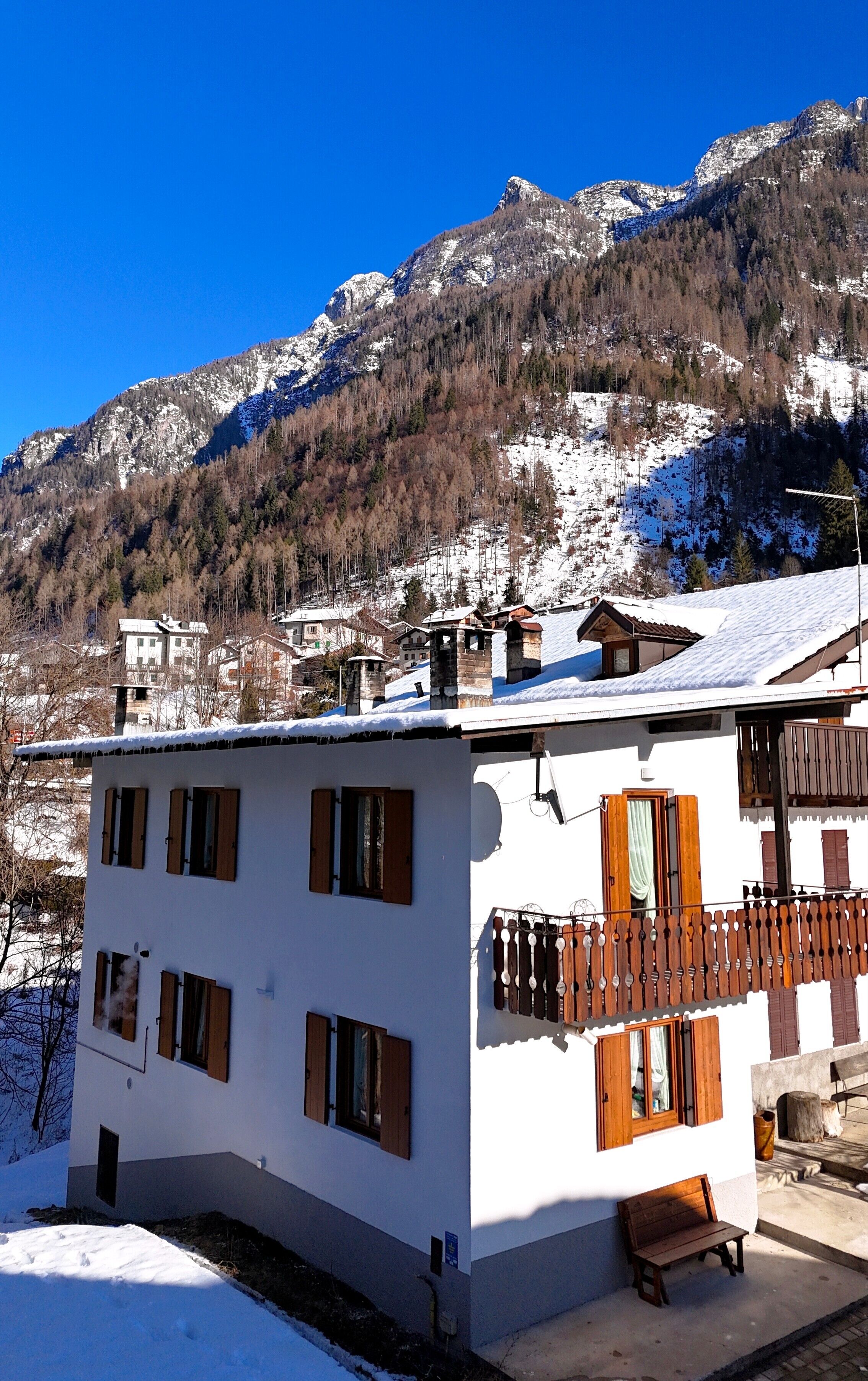 Un edificio blanco con contraventanas de madera y un balcón, con montañas nevadas al fondo.