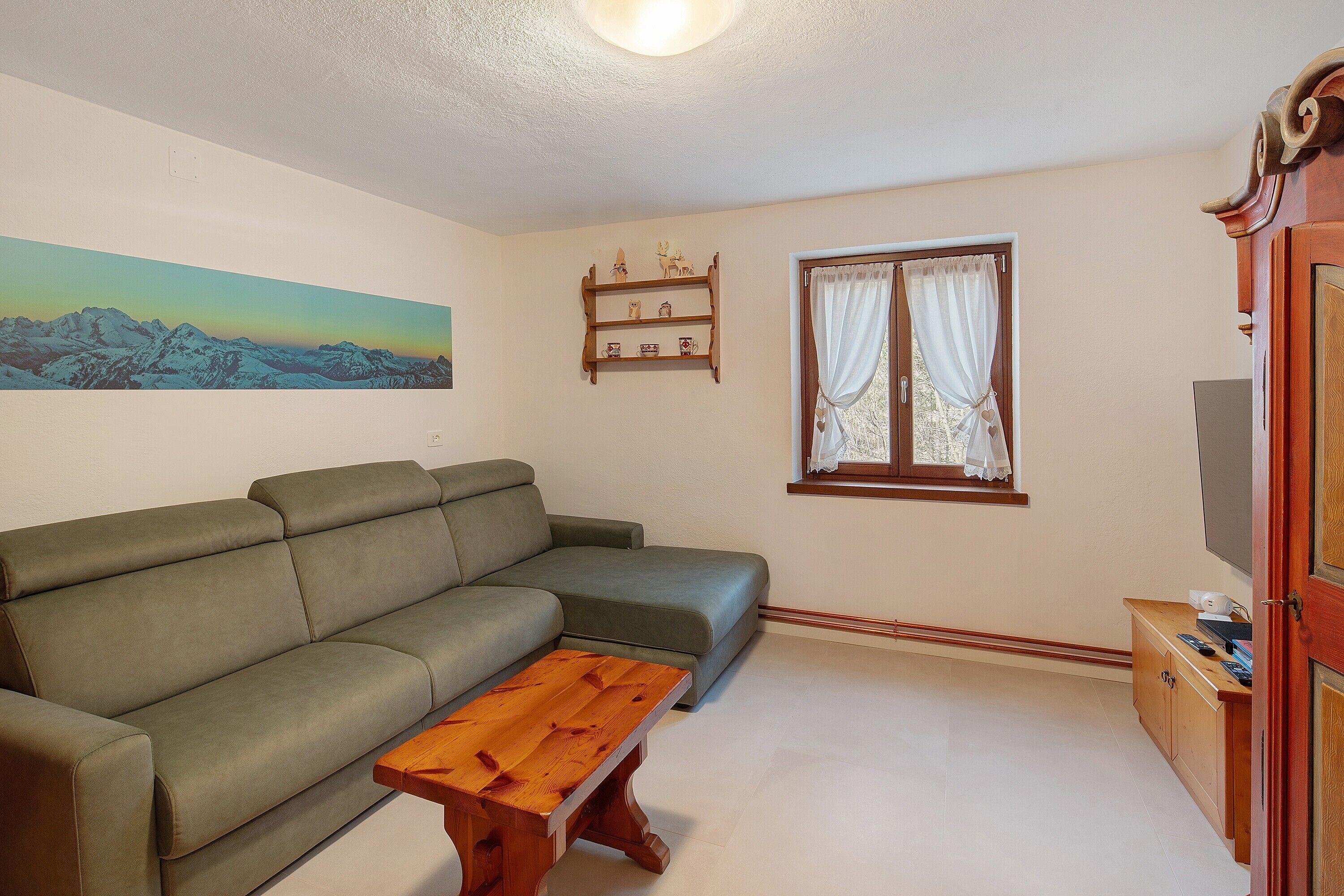 A living room with a sofa, a wooden coffee table, a painting of mountains, a window with curtains, and a TV stand.