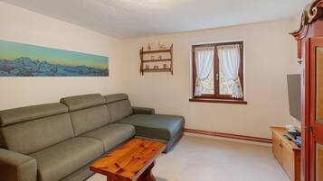 Una sala de estar con un sofá, una mesa de centro de madera, un cuadro de montañas, una ventana con cortinas y un mueble para televisión.