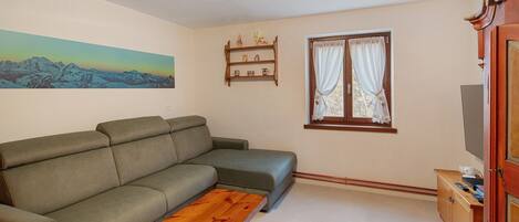 A living room with a sofa, a wooden coffee table, a painting of mountains, a window with curtains, and a TV stand.