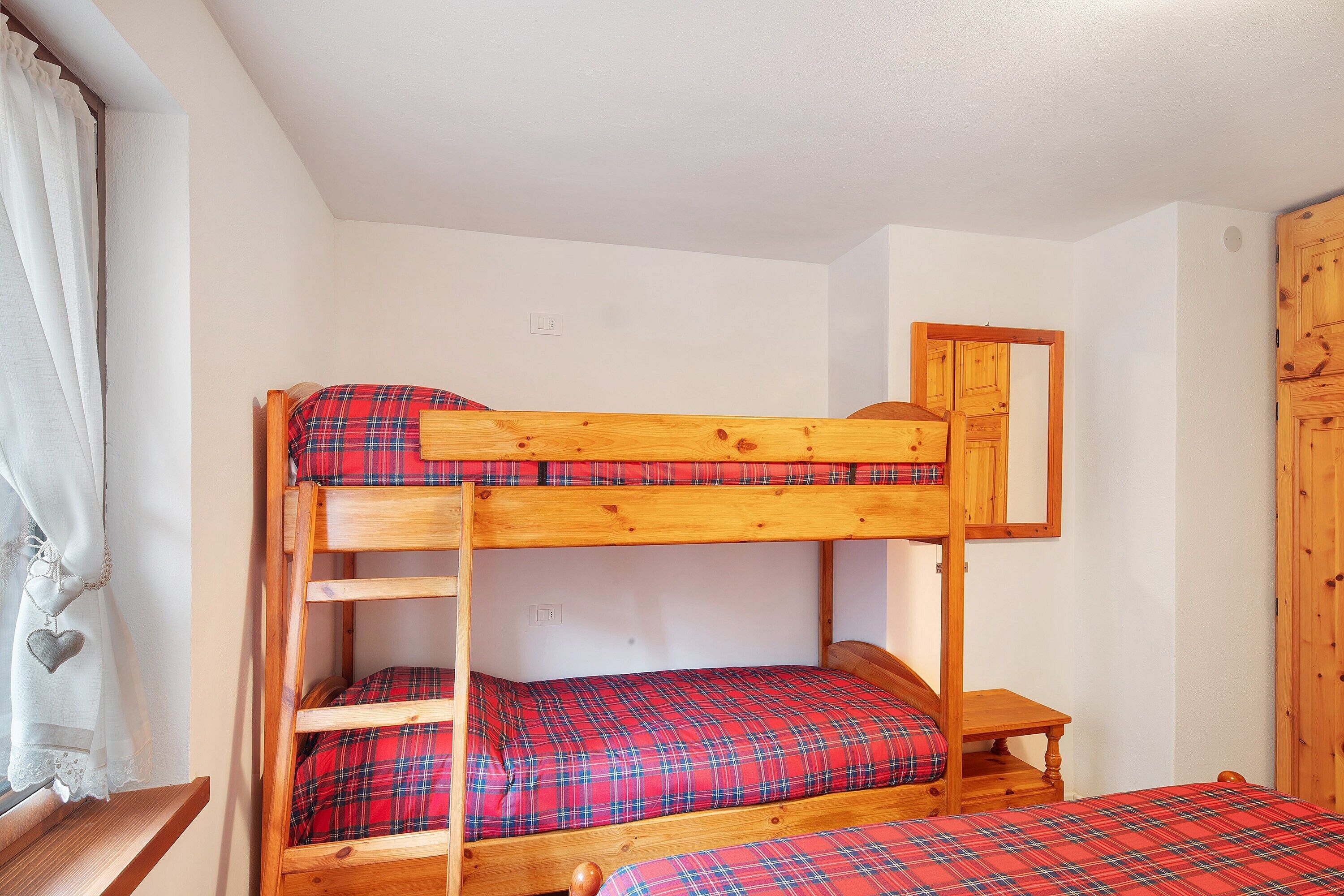 Un lit superposé avec une couette à carreaux rouges, une table de chevet en bois et une fenêtre avec des rideaux.