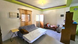 A bedroom with a bed, a small table, a chair, a window with curtains, an air conditioner, and a door.