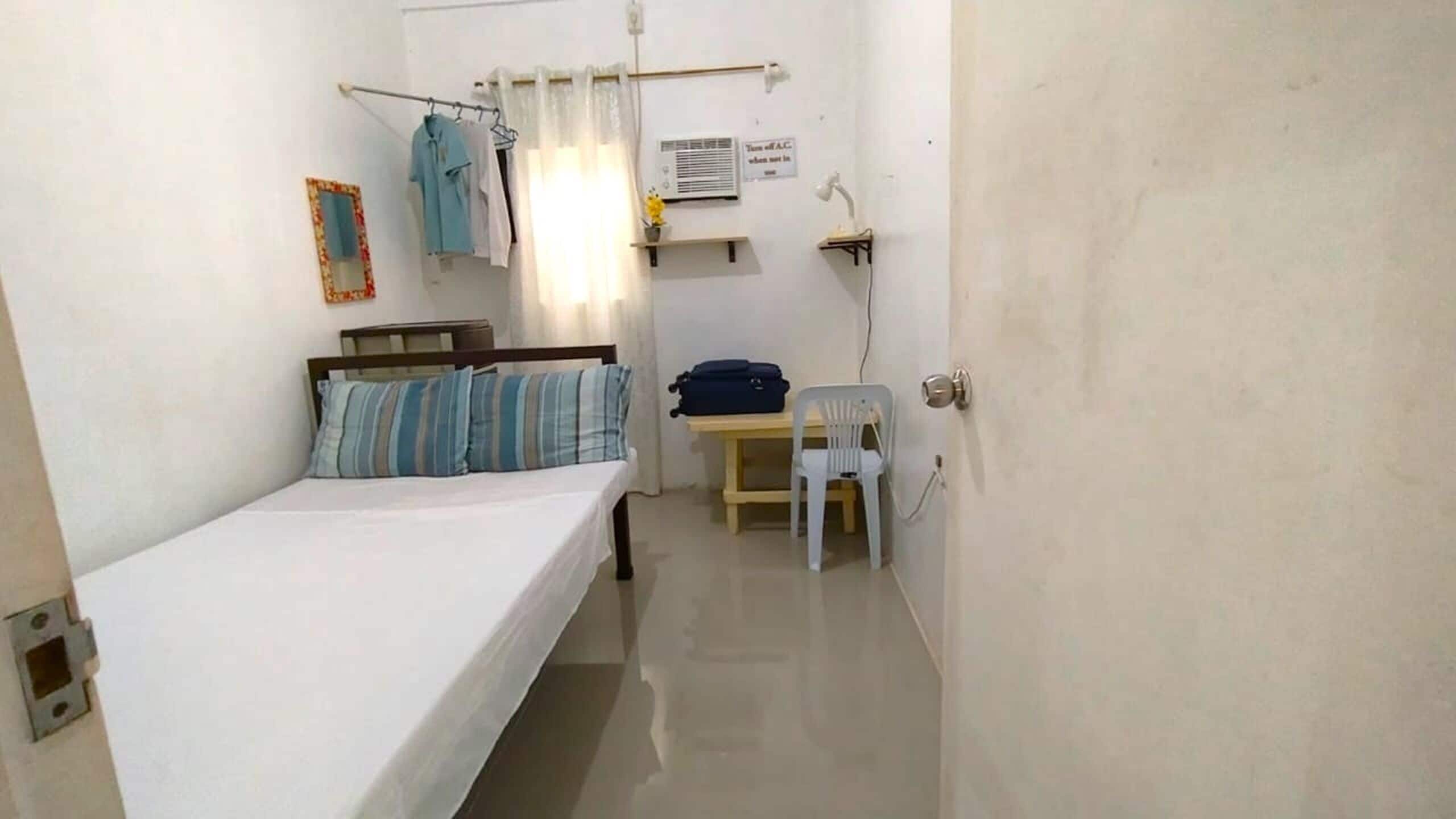 A small, narrow room with a single bed, a chair, a table, and a wall-mounted air conditioning unit.