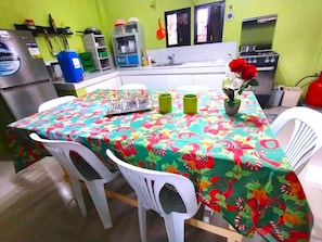 A kitchen with a table covered in a floral tablecloth, two white chairs, a refrigerator, a stove, and various kitchen items.