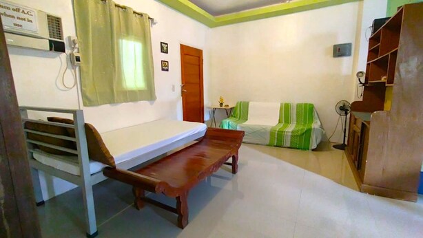 A small, simple room with a bed, a bench, a sofa, and a fan.