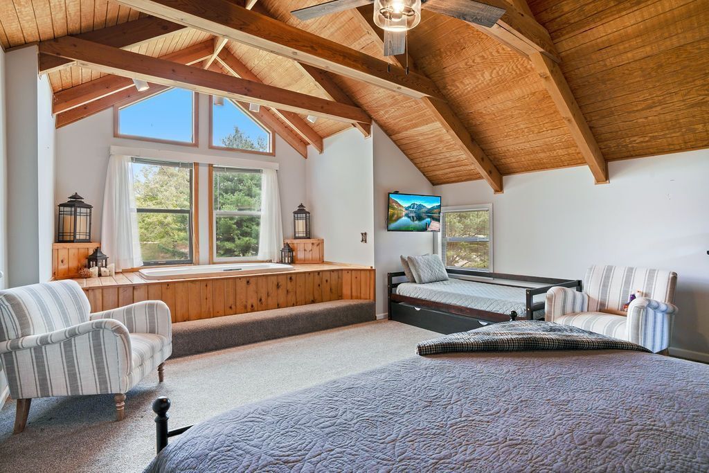 A bedroom with a large bed, a window seat, a television, and a wooden ceiling.