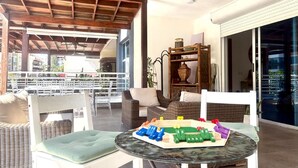 Un área de estar exterior techada con mobiliario de mimbre y un set de juguetes de madera para niños sobre una mesa de vidrio.