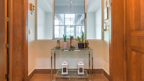 Un pasillo con una mesa consola de vidrio que exhibe estatuas religiosas y plantas, una puerta de madera y una ventana con vistas al exterior.