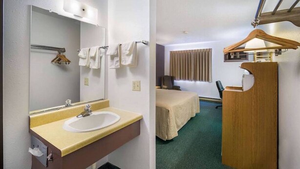 A hotel room with a bathroom, a bed, a desk, and a hanging clothes rack. - The Roosevelt Hotel & RV Park Gateway to Theodore Roosevelt National Park-Medora-on i-94 (Wibaux)