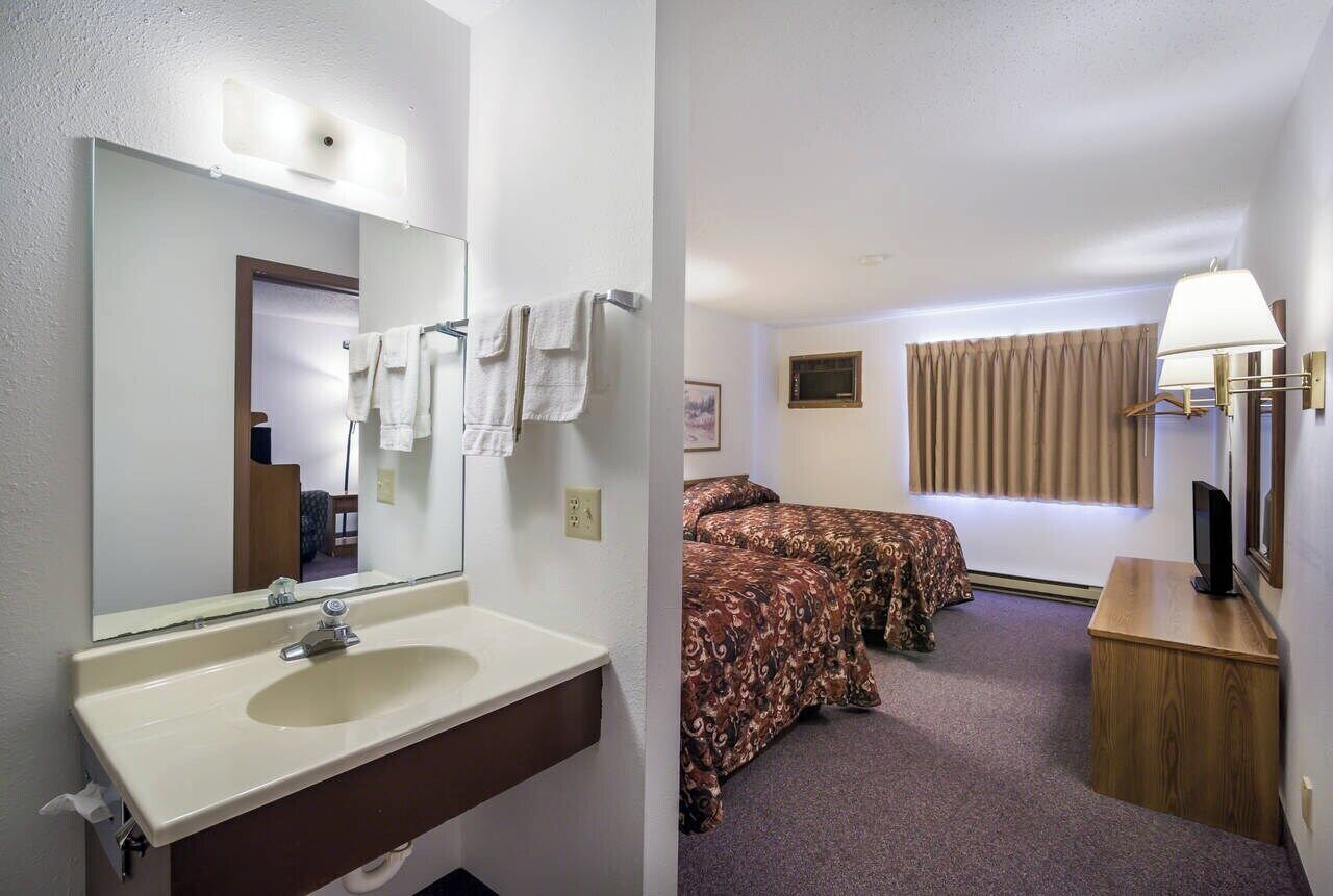 A hotel room with a bathroom featuring a sink and mirror, two beds with patterned bedding, a television, and a lamp.