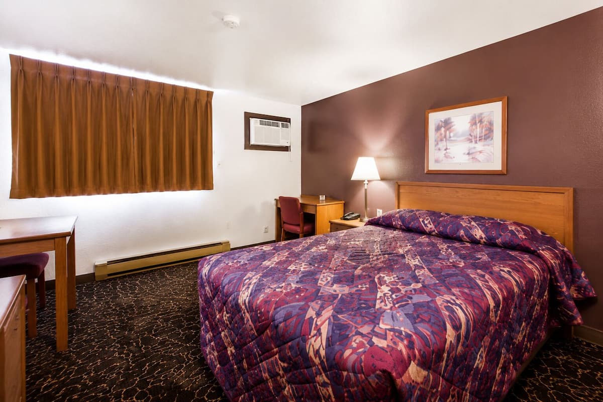 Ein Hotelzimmer mit einem Bett, einem Schreibtisch, einem Sessel, einer Lampe, einer Klimaanlage und einem gerahmten Bild an der Wand.