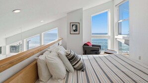 Ett sovrum med en stor säng, ett skrivbord och en stol, med utsikt över havet genom stora fönster.