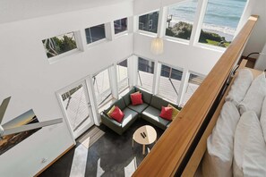 A high-ceiling room with a large window overlooking the ocean, a sectional sofa with red cushions, and a small round table. - The Lightbox | Ocean View Getaway (Tillamook)