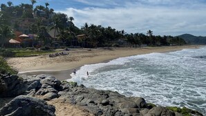 Una spiaggia con un promontorio roccioso in primo piano, palme e edifici sullo sfondo.
