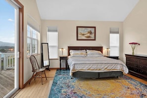 A bedroom with a large bed, a chair, a desk, and a window overlooking a deck.
