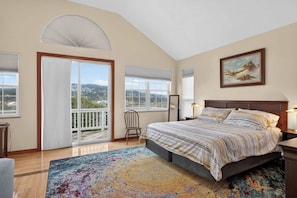 A bedroom with a large bed, a view through a sliding glass door, a painting on the wall, and a wooden floor.