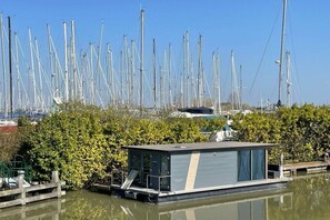A waterway with a row of tall, narrow boats, a small docked houseboat, and dense foliage in the foreground.