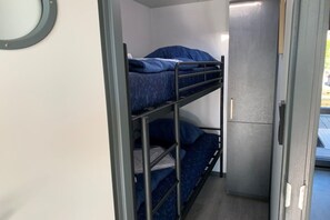 Kerrossänky, jossa on metallirunko ja siniset vuodevaatteet, pieni kaappi ja ovi, joka johtaa ulkotilaan.