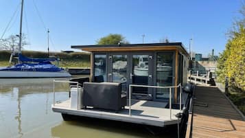 A modern floating structure with a glass enclosure, a deck with seating, and a dock extending into calm water.