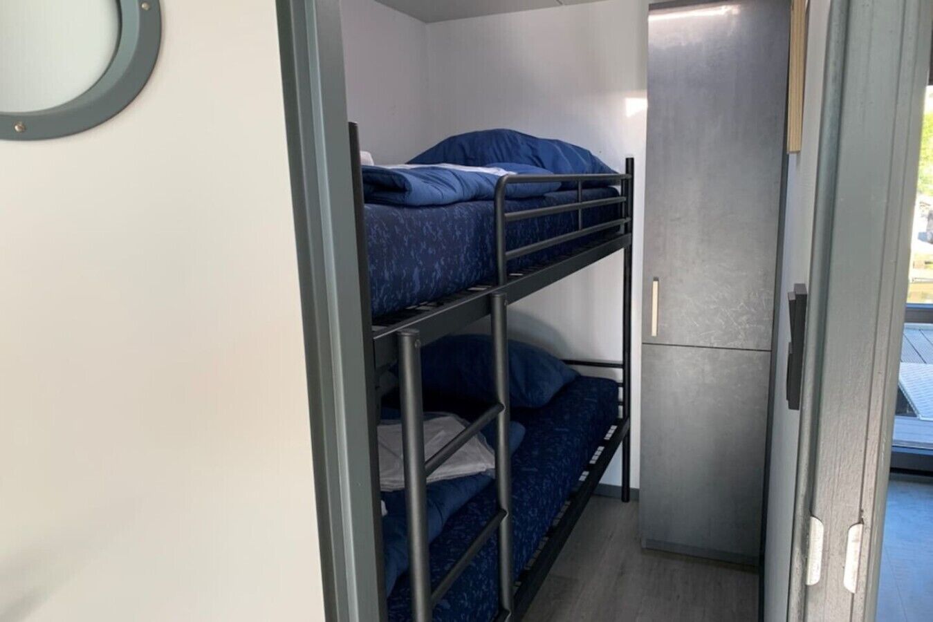 Ein Stockbett mit Metallgestell und blauer Bettwäsche, ein kleiner Schrank und eine Tür zu einem Außenbereich.