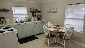 Una cocina con estufa, lavamanos y mesa de comedor con sillas. Hay dos ventanas con persianas, una estantería con utensilios de cocina y una pequeña planta en la mesa.